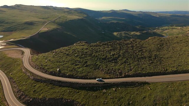 Aerial footage of a white car navigating the winding curves of Beartooth Highway in Montana during golden hour with vast alpine tundra and mountain valleys stretching into the distant horizon.