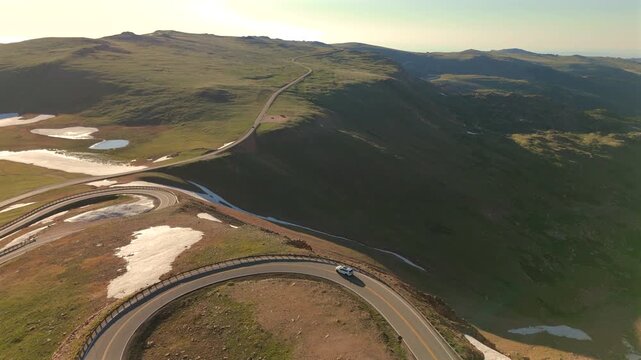 Aerial footage captures Beartooth Highway in Montana as a white vehicle navigates dramatic switchbacks through alpine terrain with snow patches and distant lake views at golden hour.