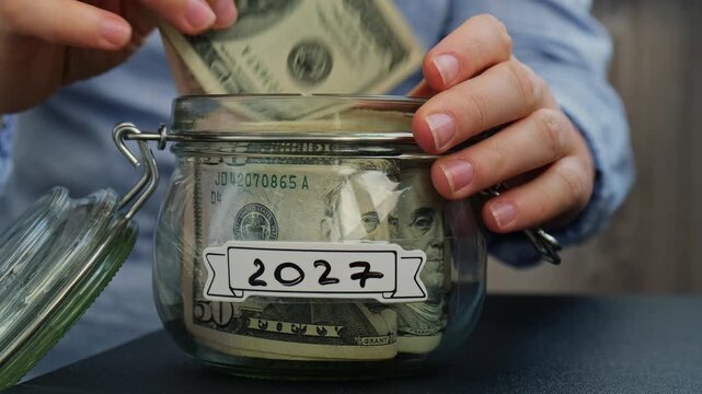 Female hands add money to Glass jar full of American currency dollars cash banknote with text 2027 year. Preparation saving money. Moderate consumption and economy. Collecting money