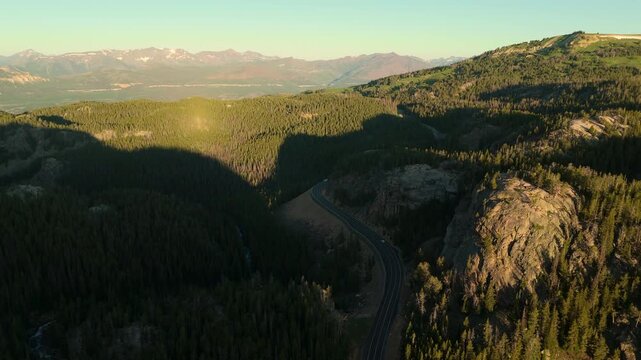 Beartooth Highway in Montana cuts through vast evergreen forests and rugged rocky cliffs during golden hour. Snow-capped peaks rise in distance as warm sunlight illuminates the scenic mountain pass.