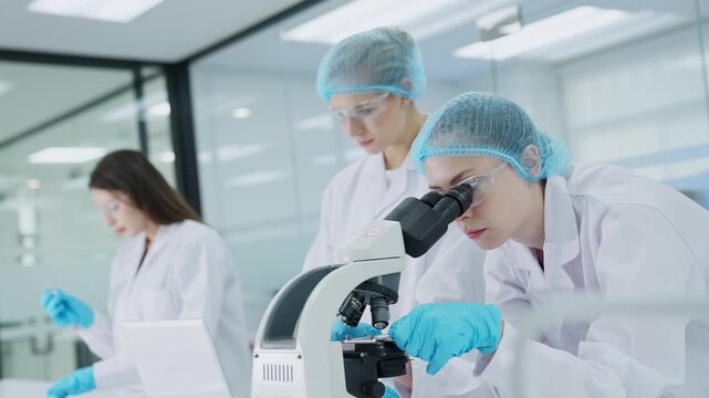 Three scientists are in a laboratory. One scientist looks through a microscope while two others observe. They conduct research on various samples and take notes on a computer.