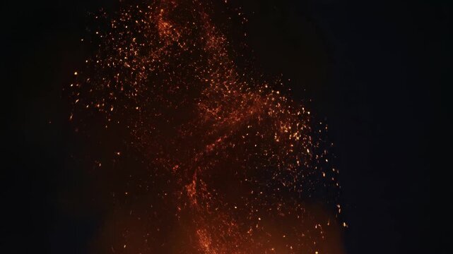 A dramatic nighttime image captures a large bonfire blazing intensely against a dark sky, sending sparks and embers into the air.