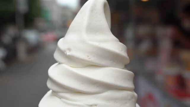 Extreme close-up shot of luxurious white vanilla soft serve ice cream with perfectly sculpted swirls held against a blurred bright street background on a warm summer day.