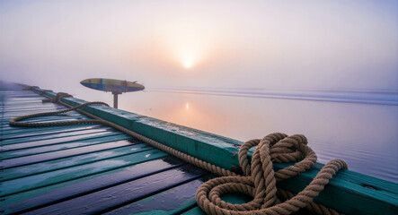 Obraz premium Nautical ropes resting on a wooden pier overlooking a misty beach at sunrise. Soft morning light illuminates the fog over calm ocean waves. Coastal serenity concept, ideal for travel marketing.