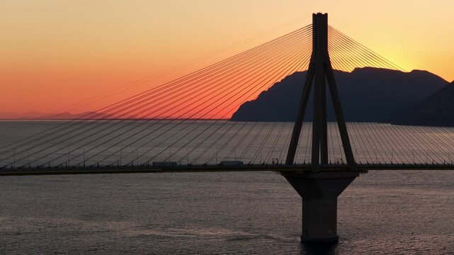 Golden sunset over the rio-antirrio bridge crossing the gulf of corinth in greece, with silhouettes of mountains and trucks driving on the charilaos trikoupis cable-stayed bridge