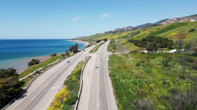 Aerial shot capturing a coastal highway and railway running parallel to the Pacific Ocean with lush hillsides. Ideal for travel documentaries or infrastructure studies.