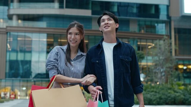 Happy young couple shopping together in city mall. Asian women and men holding colorful paper bags, walking outdoors in urban shopping district with smiling faces and modern lifestyle concept