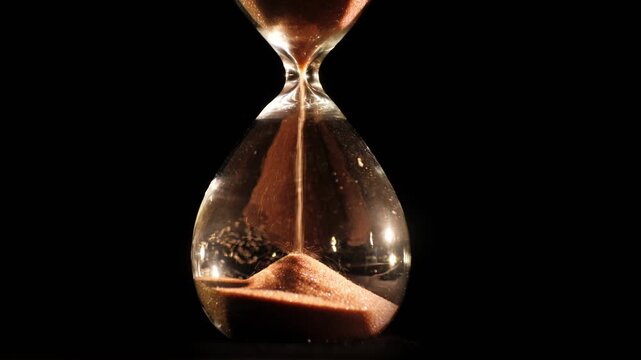 Close up slow motion of sand flowing through an hourglass, symbolizing the passage of time against a dark background with reflections.