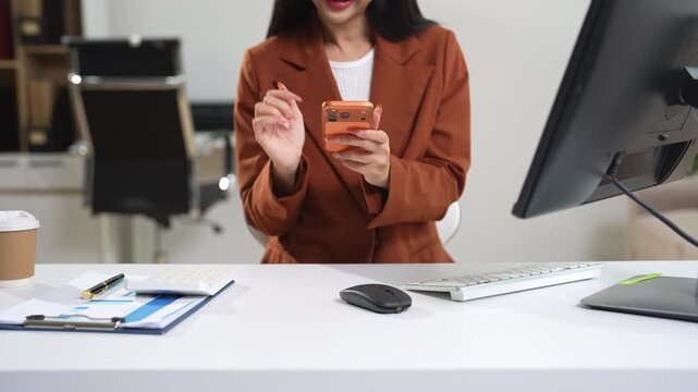 Employee, taking notes sitting at desk using smartphone and laptop searching for information at work.