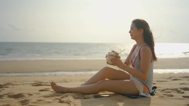 Beautiful young Asian woman relaxes on a sandy beach, using her smartphone before drinking fresh coconut water from a straw while enjoying a peaceful summer vacation looking at the ocean view.