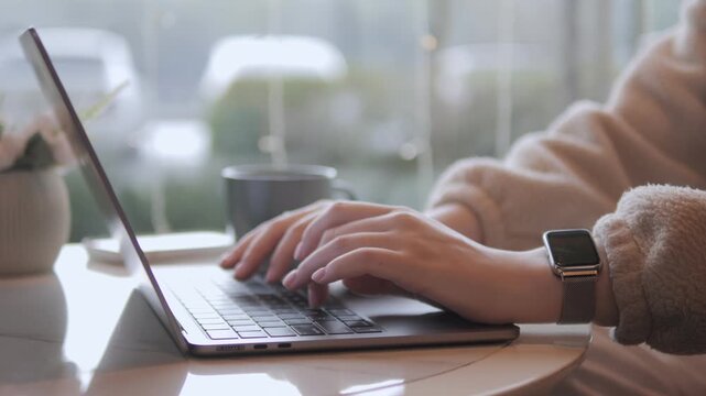 Person types on laptop near window. Mug sits beside laptop. Watch glows softly on wrist. Natural light fills room. Blurred background shows street scene. Calm workspace moment captured