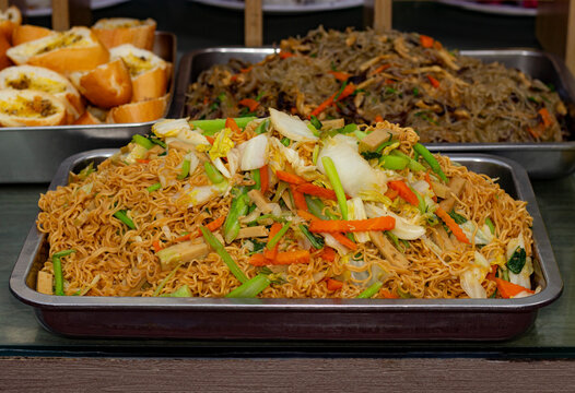 A large portion of vegan stir-fried instant noodles with mixed vegetables and plant-based protein served in a stainless steel buffet tray alongside other catering dishes.
