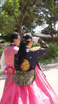Two women in traditional Korean attire walk side by side. One wears a pink hanbok with gold embroidery. The other wears a dark hanbok with floral patterns. They share a moment under leafy trees