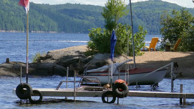 Docked small sailboat and passing powered watercraft pulling an inflatable raft on the rocky shore of Lac Simon, a recreational lake in Duhamel, Quebec, Canada.