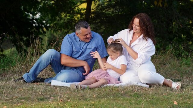 Happy mature family of three sits on blanket in summer park at sunset, laughing. Dad tickles daughter while Mom does her hair, then everyone tickles Mom. Family bonds, late fatherhood, recreation