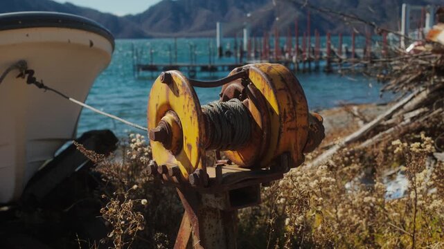 Close up of a rusty winch near a boat by the water with mountains and a pier in the background, creating a coastal industrial atmosphere.