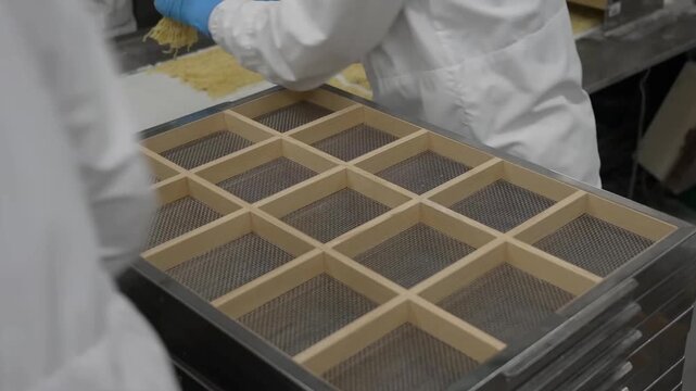 Factory workers wearing protective gear handling trays filled with fresh yellow noodles at food production facility