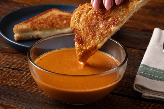 A lifestyle shot of a woman's hand dipping a grilled cheese sandwich into a tomato soup bowl
