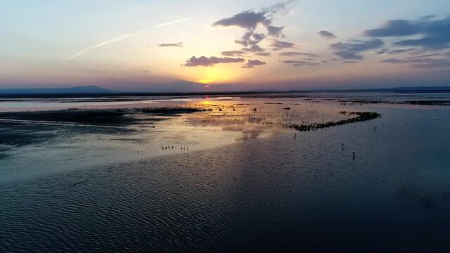 Drone flies low over wetland at sunset with perfect mirror-like reflection and dense reed patches with birds silhouetted against glowing sky in Iranian marsh footage.