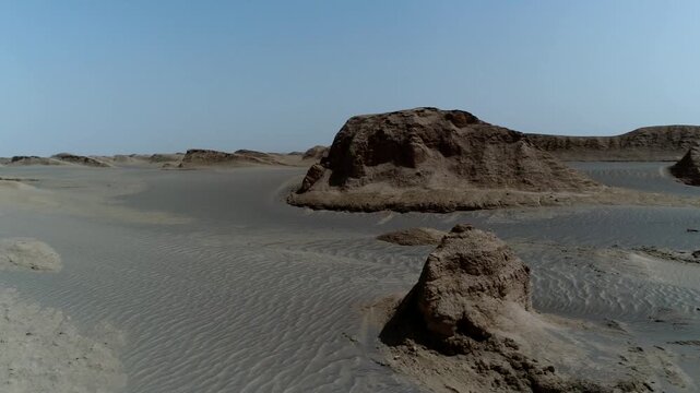 Final aerial perspective shows wind-sculpted rock formation rising from flat desert with distant dunes under bright blue sky in central Iran stock video production.