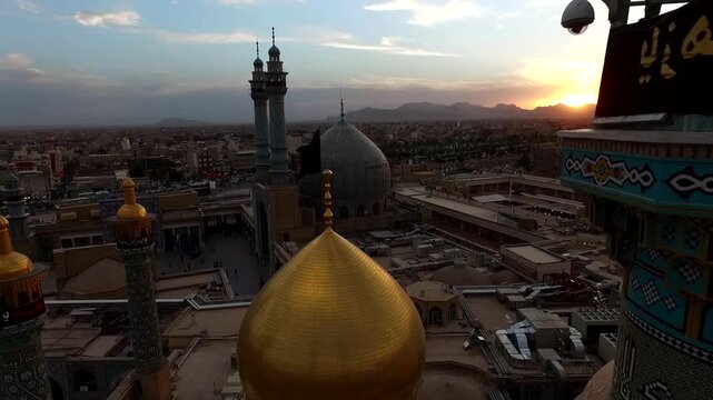 High altitude aerial captures full shrine skyline with golden dome and multiple minarets surrounded by old Qom city at sunset with pilgrims visible below Iran stock footage.