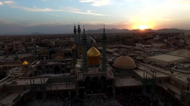 Wide aerial reveals Lady Masoumeh shrine with golden dome and four minarets at sunset surrounded by old city rooftops and pilgrims in Qom Iran stock footage.