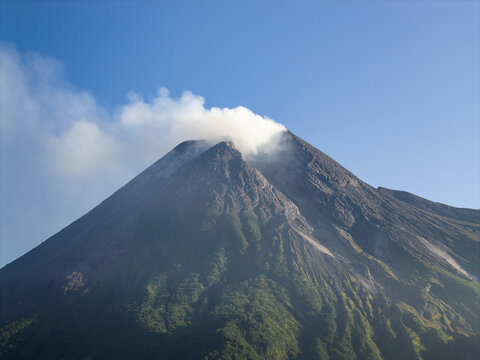 Aerial view of Mount Merapi with smoke rising from the crater and lush green slopes under a clear blue sky Kecamatan Pakem, Daerah Istimewa Yogya, Indonesia.