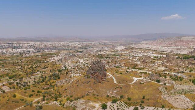 Goreme, Nevsehir, Turkey. A towering volcanic tuff rock formation rises from the golden Cappadocia plateau, aerial panoramic view over eroded valleys and distant mountain ridges under clear summer sk