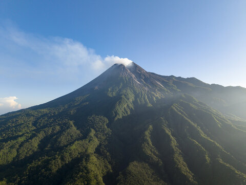 Aerial view of Mount Merapi volcano with smoke rising from the crater and lush green forested slopes under a clear blue sky Kecamatan Pakem, Daerah Istimewa Yogya, Indonesia.