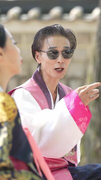Woman in vibrant hanbok gestures animatedly. She wears sunglasses and speaks with intensity. Another woman listens, partially turned away. Scene suggests a playful or confrontational dialogue