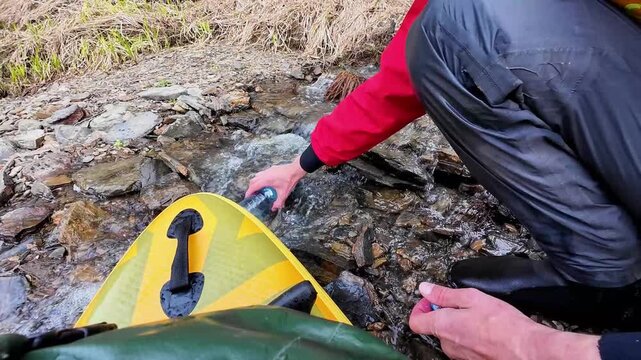 Kayak refill from mountain stream rocks, camper reaches to scoop fresh water into bottle beside yellow kayak on shallow creek, POV closeup showing hands, hull and pebble bank, practical