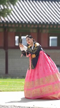 Woman in traditional dress stands gracefully. She holds a book, gazing thoughtfully upward. Ancient roofline frames the serene moment. Soft daylight highlights her colorful gown