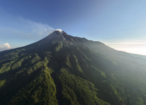 Aerial view of Mount Merapi volcano with steam rising from the crater above lush green forested slopes and Bunker Kaliadem in Kecamatan Pakem, Daerah Istimewa Yogya, Indonesia.