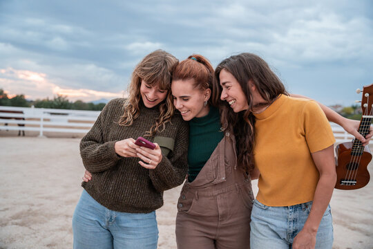 Three friends laugh while looking at a phone outdoors