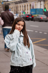 Fototapeta premium Young Girl Enjoying a Moment of Contemplation on a Bustling City Street at Dusk. In a lively urban setting, a girl stands thoughtfully on the sidewalk, wearing a light denim jacket