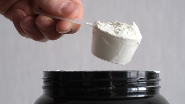 Male hand holds measuring spoon with brown protein on gray background side view.