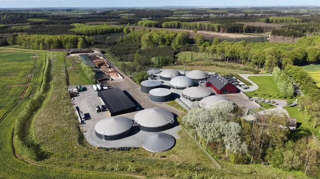 Sweeping aerial view of a complete biogas facility with numerous digester tanks surrounded by forests and agricultural fields in Hammel, Denmark.
