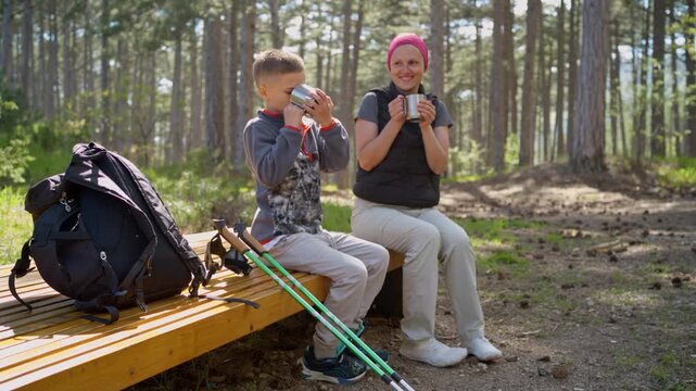 Mother and son drinking tea during forest hike. Happy family sits on wooden bench in pine forest during mountain trip. 