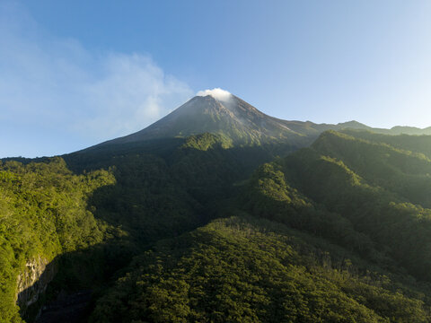 Aerial view of Mount Merapi volcano with a smoking peak and lush green forested slopes under a clear blue sky in Kecamatan Pakem, Daerah Istimewa Yogya, Indonesia.