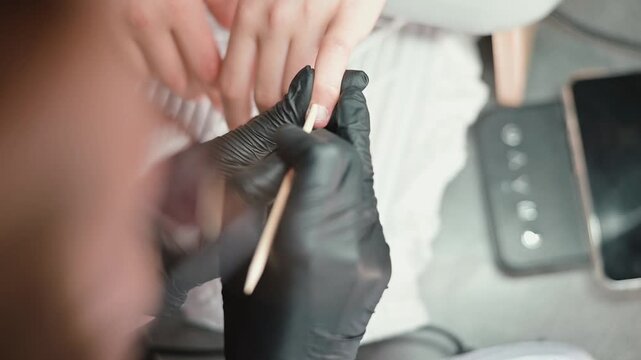 A person with gloves applies nail treatment while another hand is prepared for care. The setting includes tools and devices commonly found in nail care environments