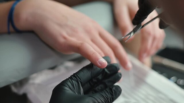 A nail technician trims and shapes the client nails before applying enhancements in a beauty studio. The technician wears black gloves while focusing on the client's hands