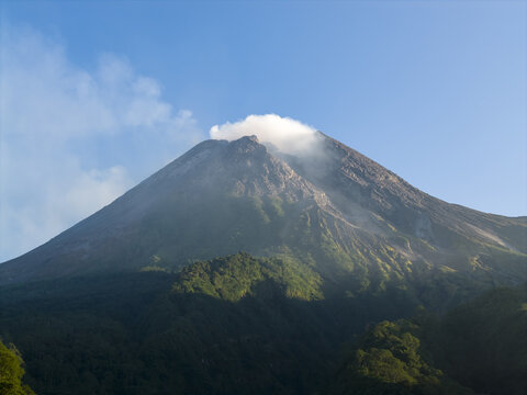 Aerial view of Mount Merapi volcano with smoke rising from its crater and lush green forests on the slopes under a clear blue sky Kecamatan Pakem, Daerah Istimewa Yogya, Indonesia.