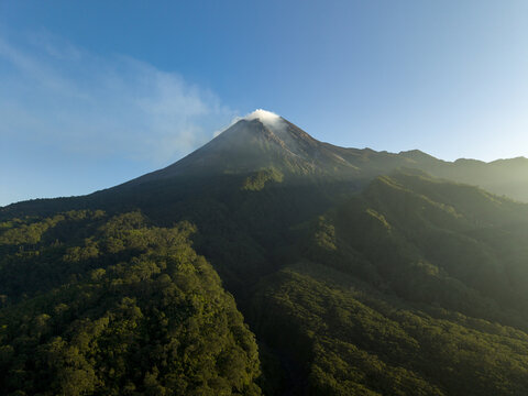 Aerial view of Mount Merapi with smoke rising from the peak and lush green forests on the slopes in Kecamatan Pakem, Daerah Istimewa Yogya, Indonesia.