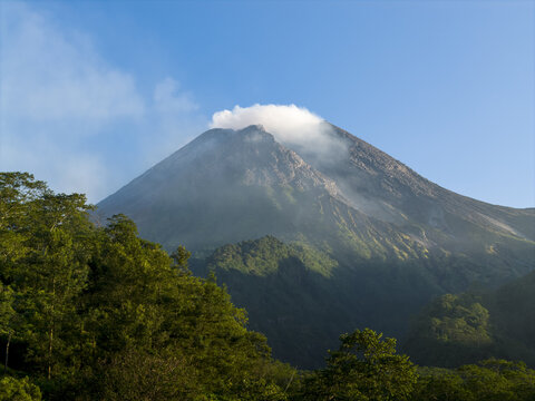 Aerial view of Mount Merapi volcano with smoke rising from the crater above lush green forests and hills in Kecamatan Pakem, Daerah Istimewa Yogya, Indonesia.