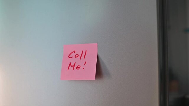 A hand reaches towards a pink sticky note on a fridge that says Call Me. This action captures the moment of responding to a reminder in a domestic setting