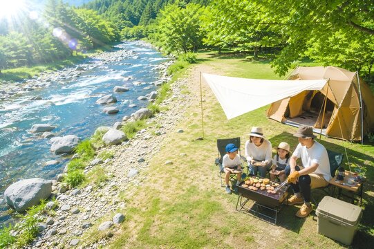 夏の強い日差しが降り注ぐ河川敷で家族が楽しそうにバーベキューをするアウトドアレジャーイメージ（AI生成）