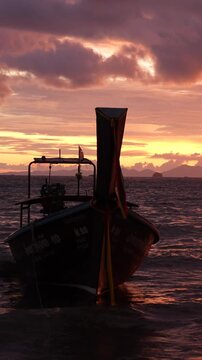 Vertical slow-motion shot of a traditional Thai long-tail boat silhouetted at sunset, floating on gentle sea water beneath dramatic glowing clouds.