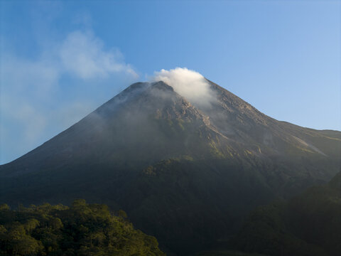 Aerial view of the smoking peak of Mount Merapi volcano with lush green slopes and a clear blue sky near Kecamatan Pakem, Daerah Istimewa Yogya, Indonesia.