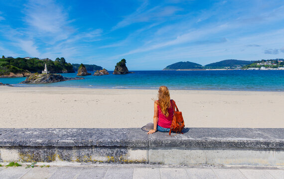 Woman with backpack sitting on promenade wall looking at os castelos rocks in viveiro, Summer vacation, tour tourism in spain