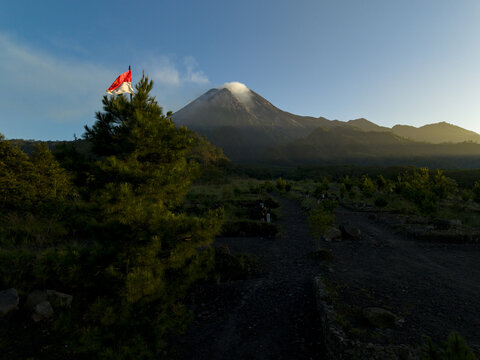 Aerial view of Bunker Kaliadem Merapi with the smoking Mount Merapi volcano, an Indonesian flag on a tree, and lush greenery in Kecamatan Pakem, Daerah Istimewa Yogya, Indonesia.